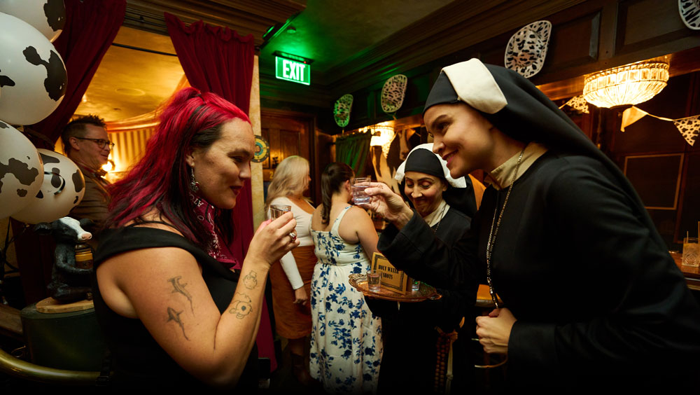 Sister Maria sharing a cocktail shot with a guest inside The Cow Hide Bar inside Atomic Saloon Show.